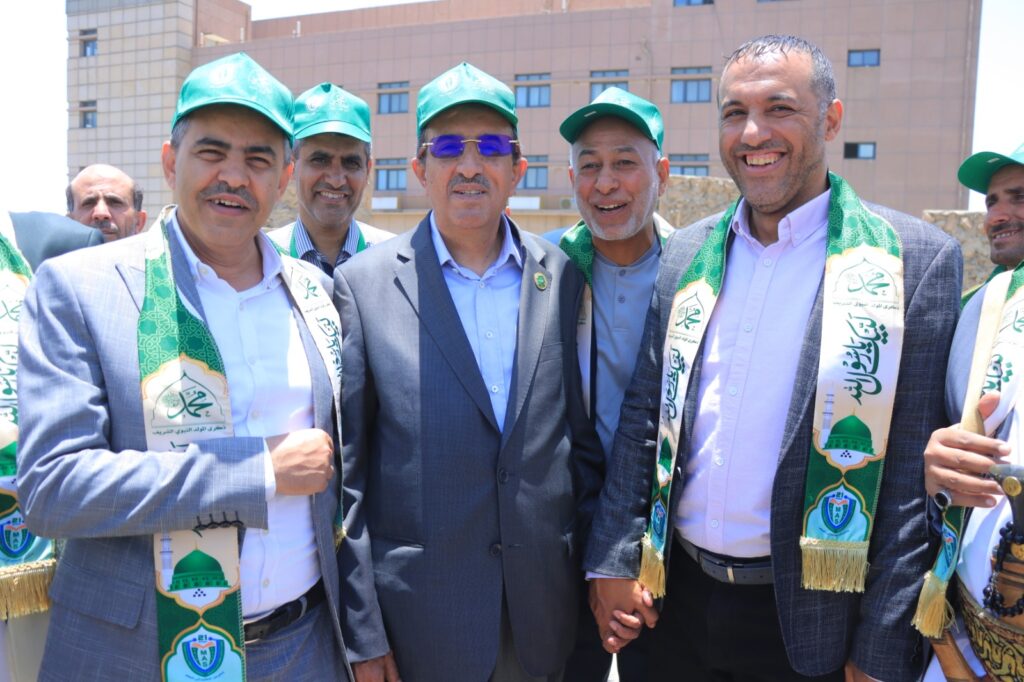 Group of men in suits and green sashes posing together outdoors at a ceremony, smiling. Caps and sashes display a mosque emblem and Arabic text.