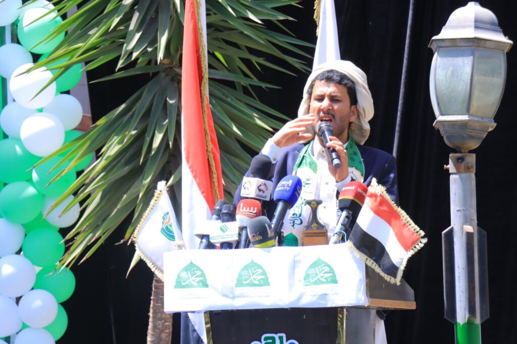 Man in a suit speaks into a handheld microphone at a decorated podium with flags and logos, outdoors with balloons nearby.