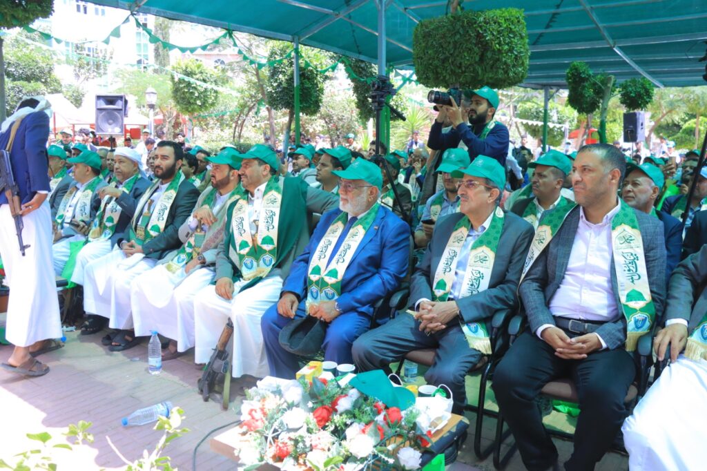 Audience of men in green caps and sashes seated under a canopy at an outdoor ceremony; a woman in blue passes by with a rifle slung over her shoulder.