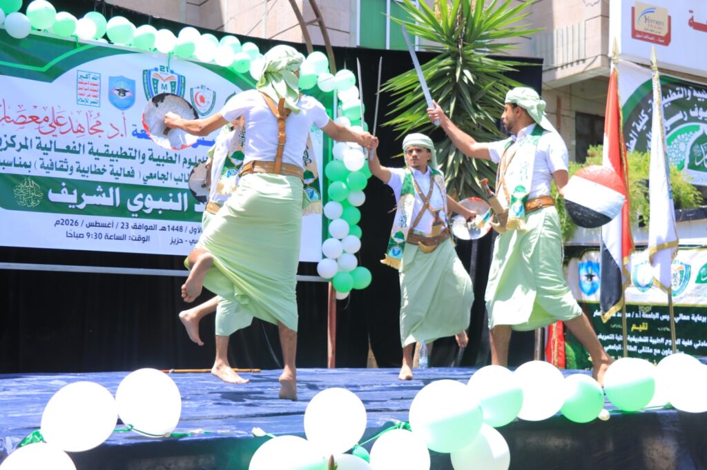 Group of men in green traditional outfits performing a sword dance on a stage, surrounded by green‑and‑white balloons and banners.