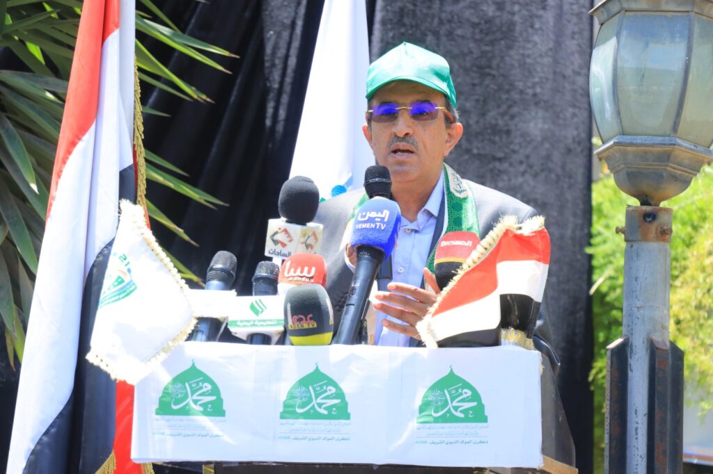 Man in a green cap and glasses speaking into multiple microphones at an outdoor podium, flanked by flags and banners.
