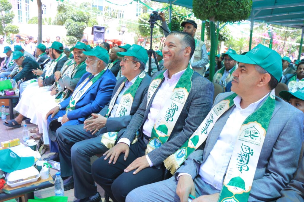 Dignitaries seated outdoors wearing green scarves and caps at a formal ceremony under a canopy.