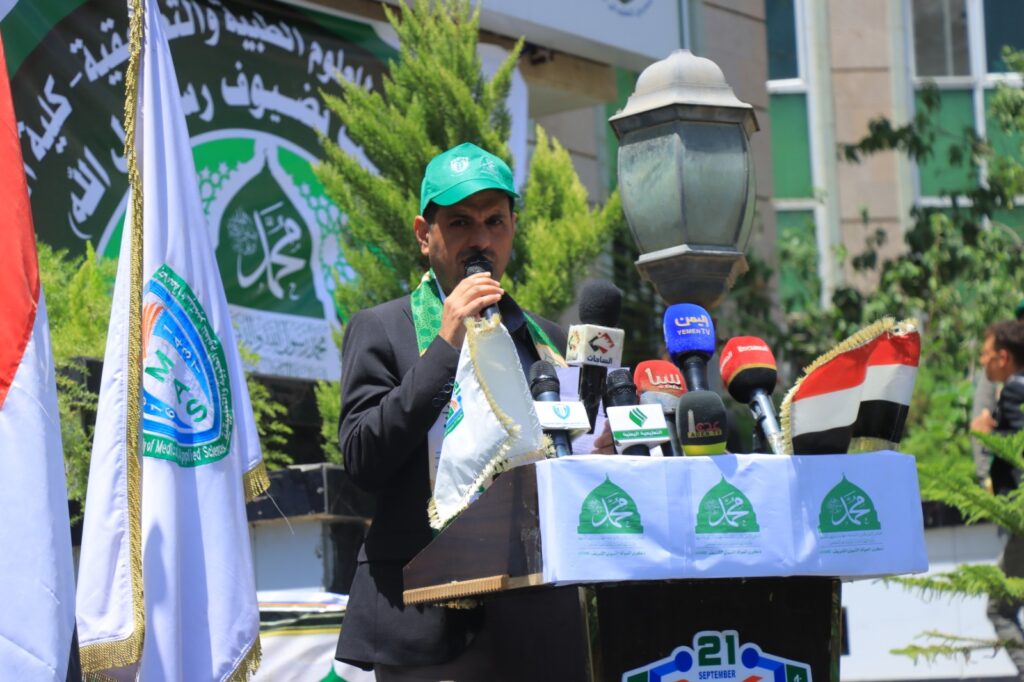 Man in a suit speaking into a microphone at a podium during an outdoor event, green cap and scarf, multiple TV microphones nearby.