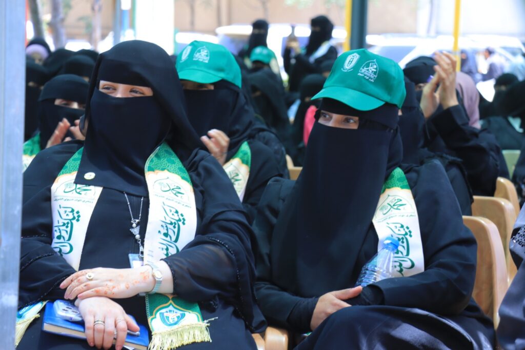 Women in black abayas and niqabs sit at an outdoor event, wearing green caps and sashes with Arabic text.