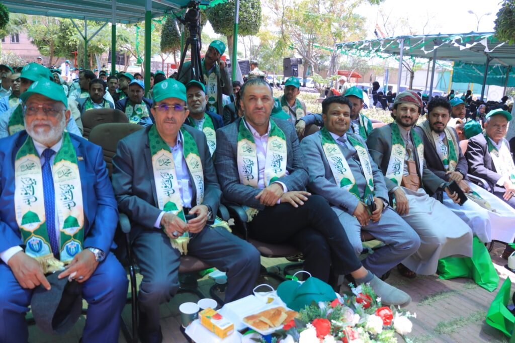 Row of men in suits with green scarves and caps seated outdoors at a ceremony, a camera on a tripod behind them.