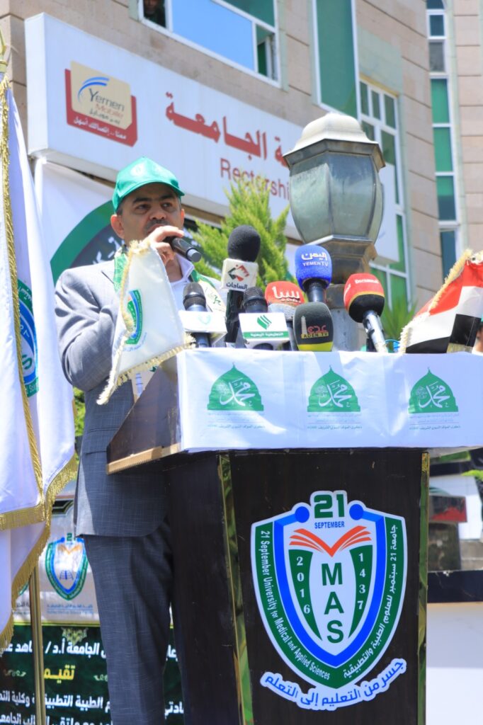 Man in a gray suit and green cap delivering a speech at a podium with multiple microphones, university banners and a MAS emblem visible on the lectern.