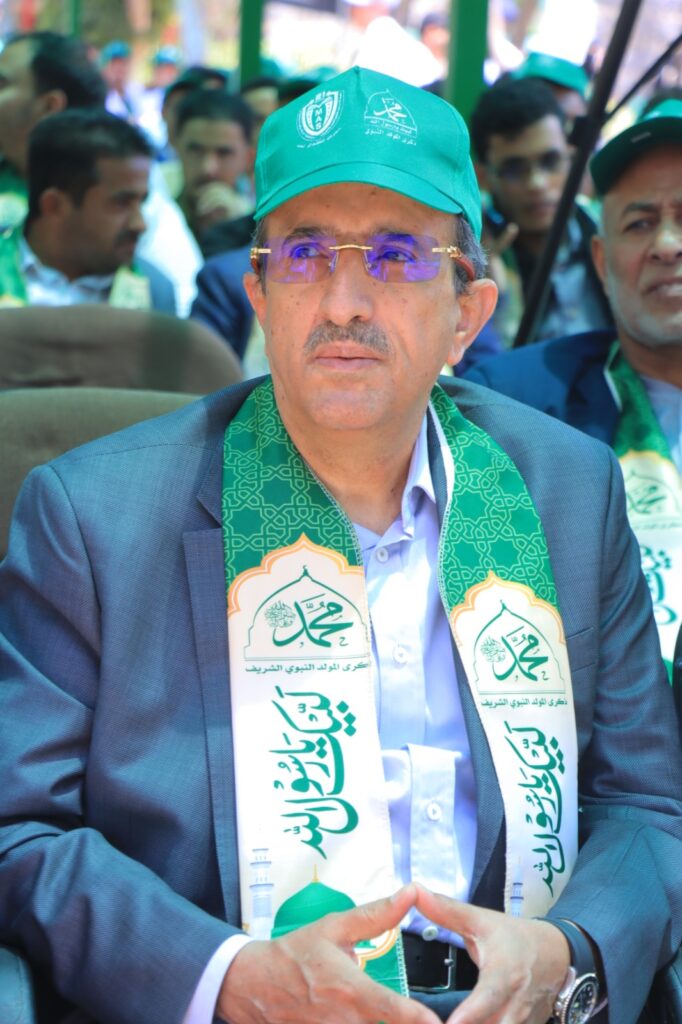 Middle-aged man in a blue suit wearing a green cap and patterned green scarf with Arabic script at an outdoor event.