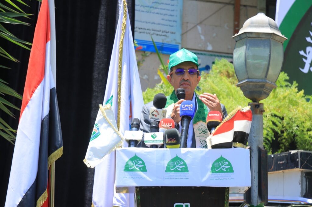 Man wearing a green cap and glasses speaks into a microphone at a podium, with multiple news microphones and flags in the background.