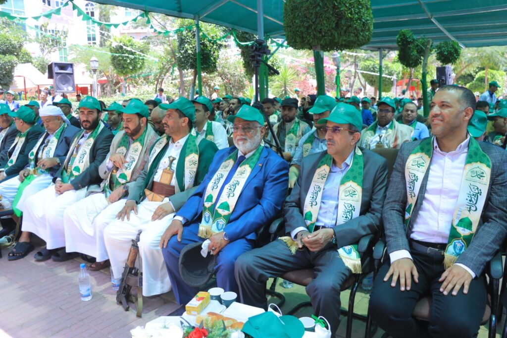 Group of men in green caps and scarves seated at an outdoor ceremony, formal attire with green decorations overhead and attendees in the background
