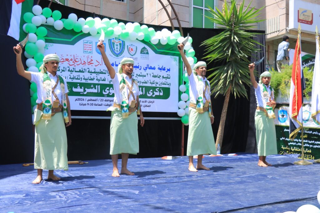 Four men in traditional Yemeni attire perform a knife dance on a blue stage with a green-and-white balloon arch and Arabic banner behind them.