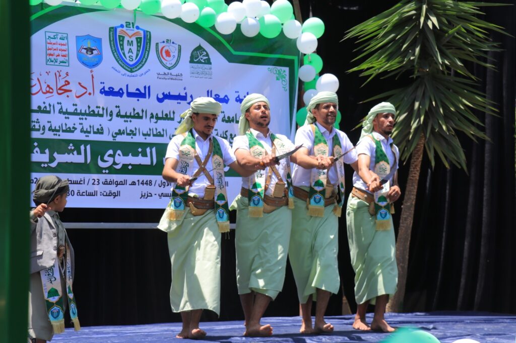Four men in green traditional outfits perform a sword dance on a stage with an Arabic banner backdrop.