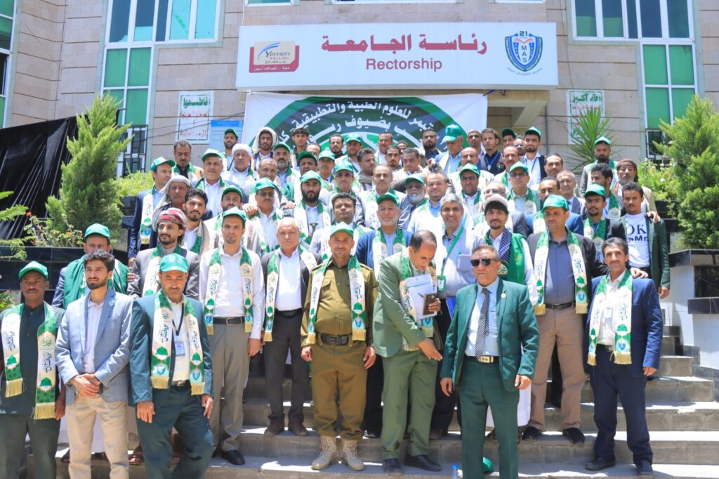 Group photo of faculty and students wearing green scarves and caps in front of a university building with a banner, posing for a ceremony at Rectorship.