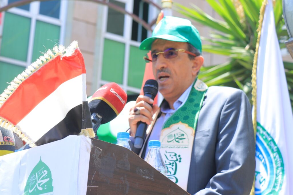 Man speaking at a podium outdoors, holding a microphone with flags and banners nearby.