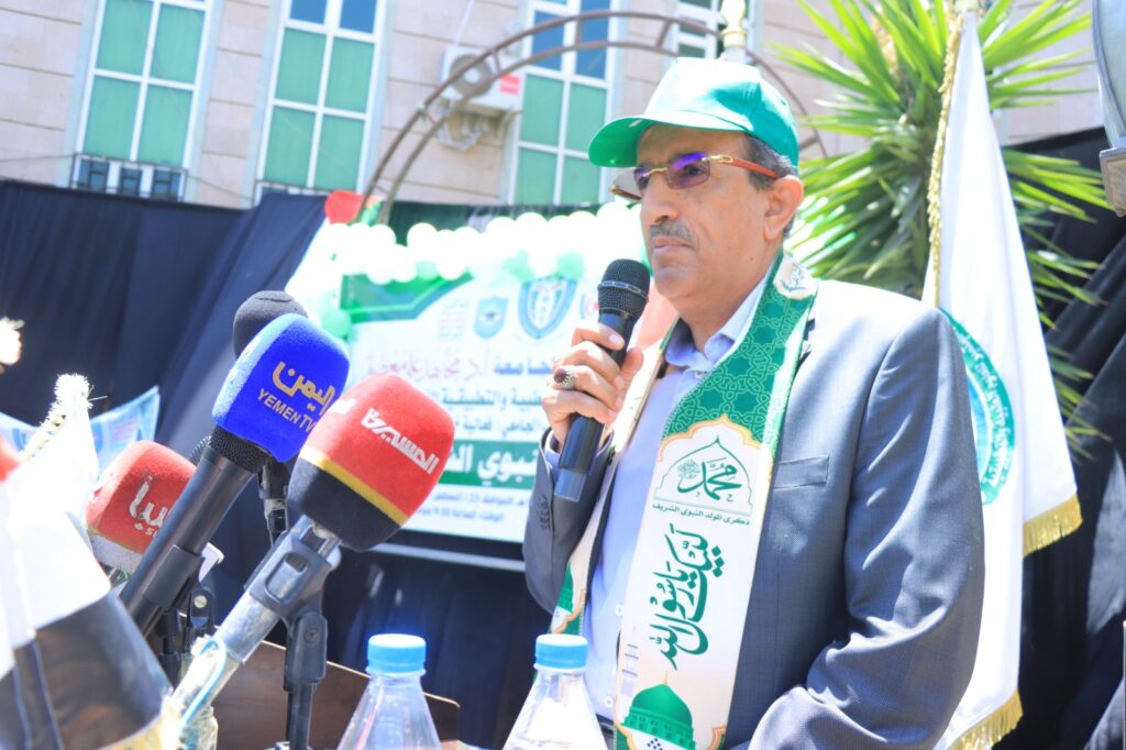 Man in suit speaking into a microphone at a press conference, wearing a green cap and scarf with an Islamic emblem, banners in the background.
