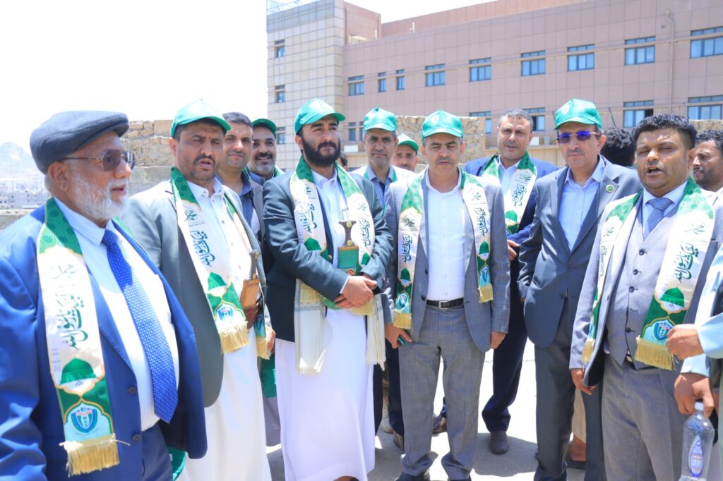 Group of men in suits and traditional robes wearing green ceremonial sashes and caps, standing together outdoors.