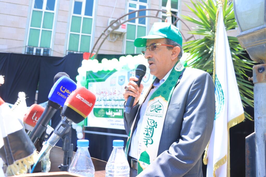 Man in a suit and green cap speaks into a handheld microphone at an outdoor press event, reporters' colorful mics in the foreground.