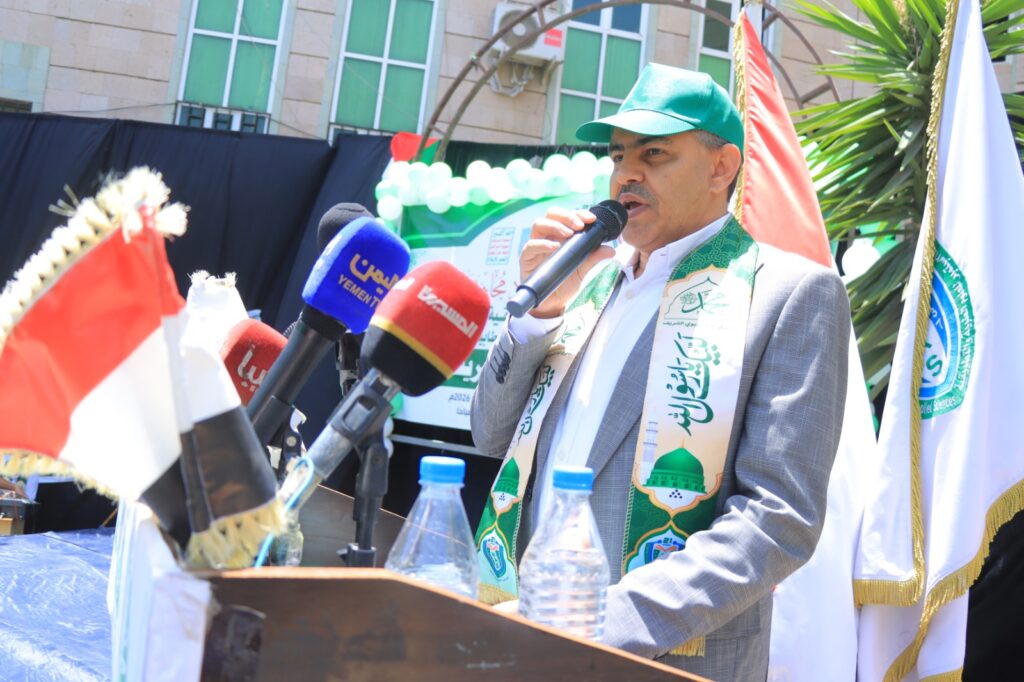 Man in a green cap and patterned scarf speaks into a handheld microphone at an outdoor podium, with flags and multiple microphones nearby.