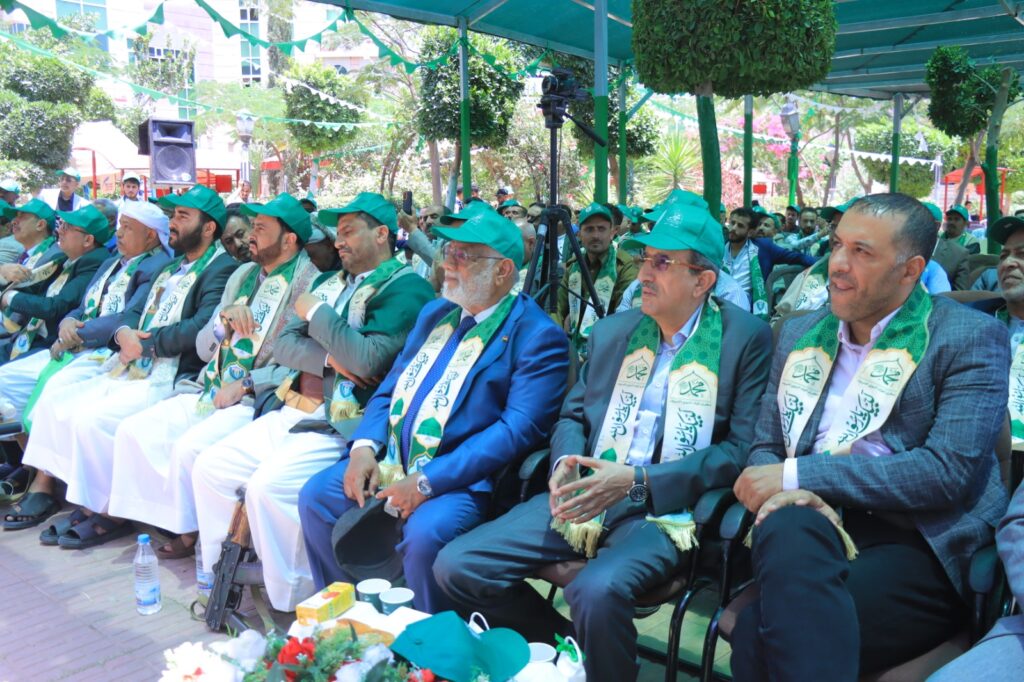 Group of men in suits wearing green caps and scarf-like sashes seated at an outdoor event under a canopy.