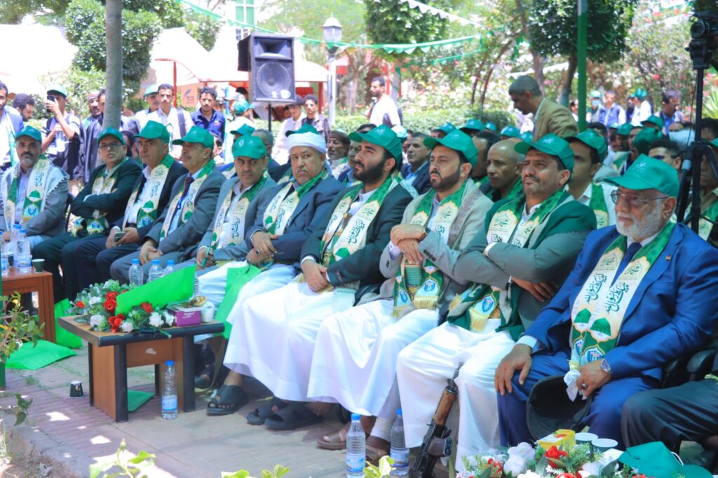 Group of men in green caps and scarves seated in a row at an outdoor ceremony, with onlookers and a speaker setup in the background.