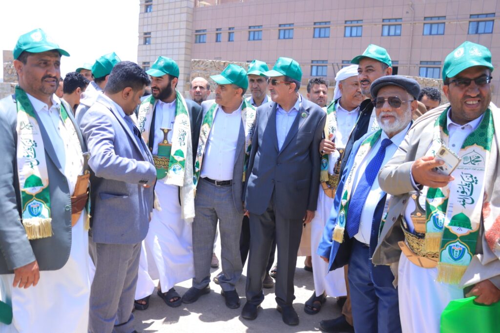 Group of men in green hats and ceremonial scarves at an outdoor gathering, shaking hands, with a building in the background.