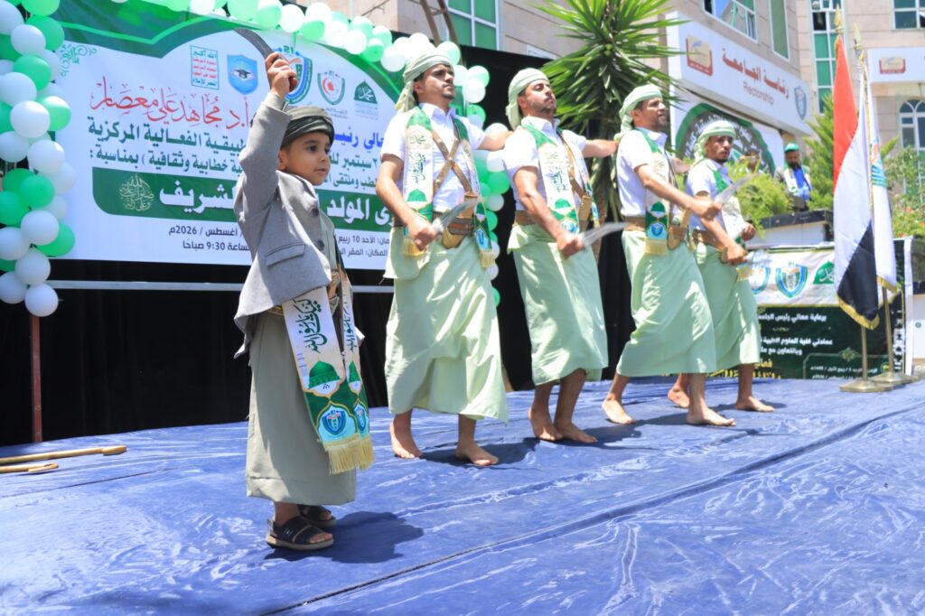 Young boy raising his arm while a line of men in traditional attire perform a stage dance at a cultural festival, with green balloons and Arabic banners in the background.