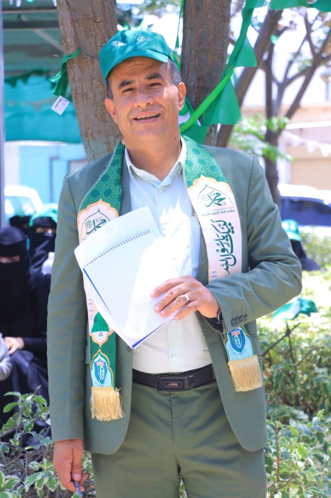 Man in a green suit and cap holding a notebook at an outdoor event, with green banners and trees in the background.