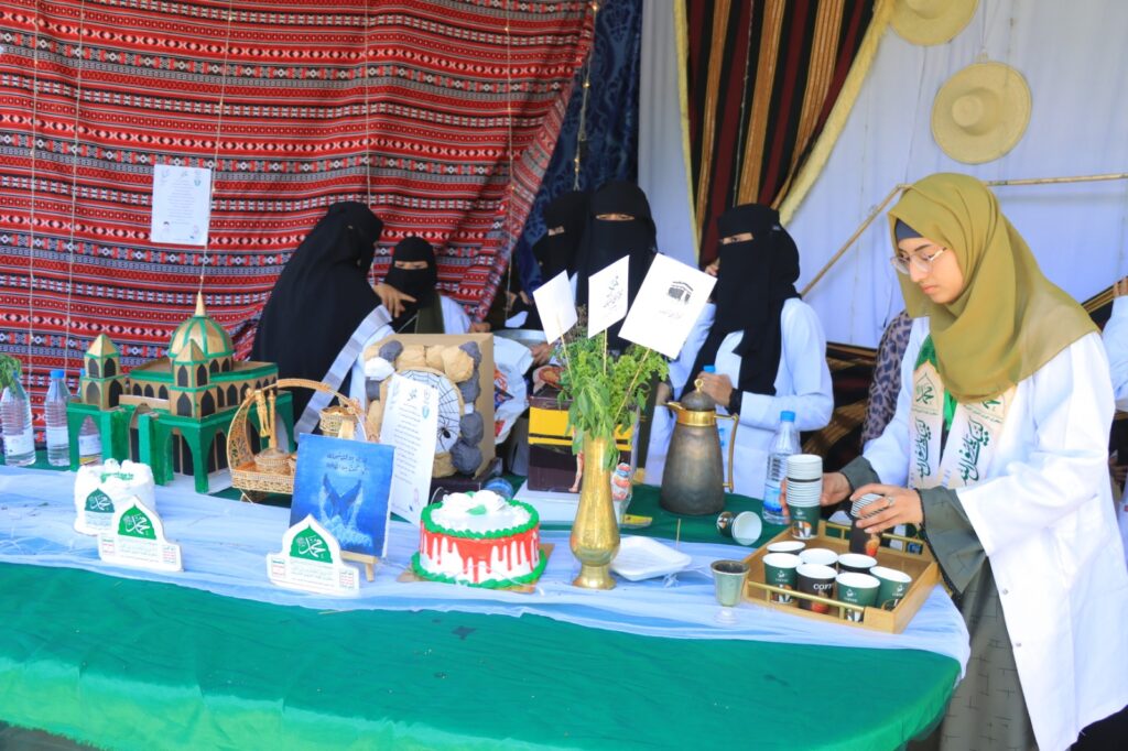 Women in traditional dress and hijabs run a cultural market stall with crafts, cakes, and beverages on a long table covered in blue and green cloths.