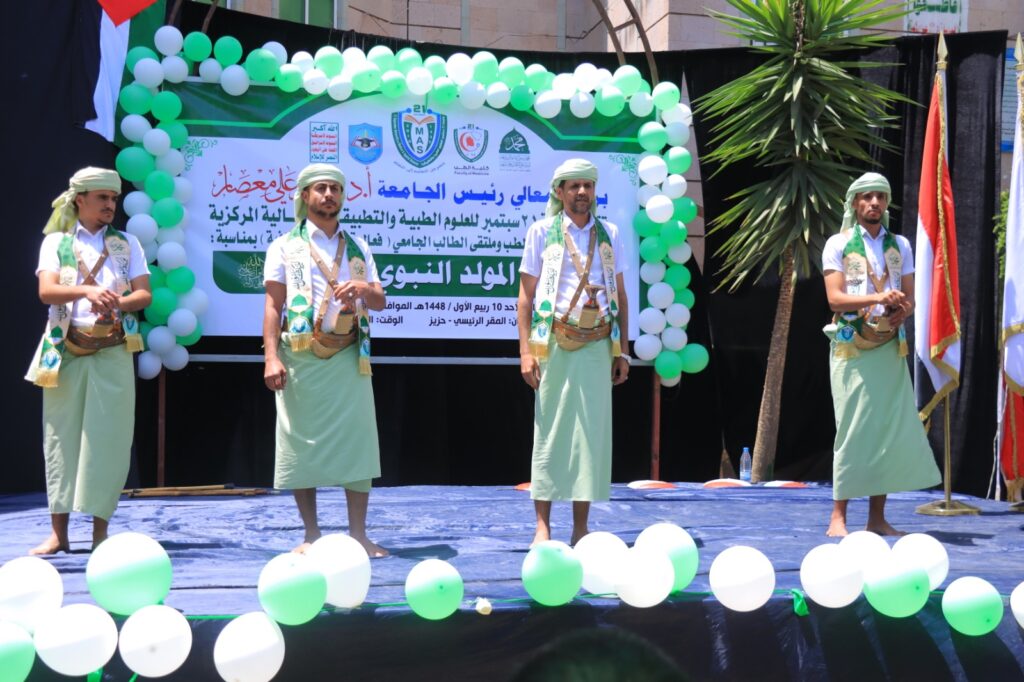 Four men in light green traditional garments stand on a blue stage under a green/white balloon arch, with a banner behind them in Arabic.