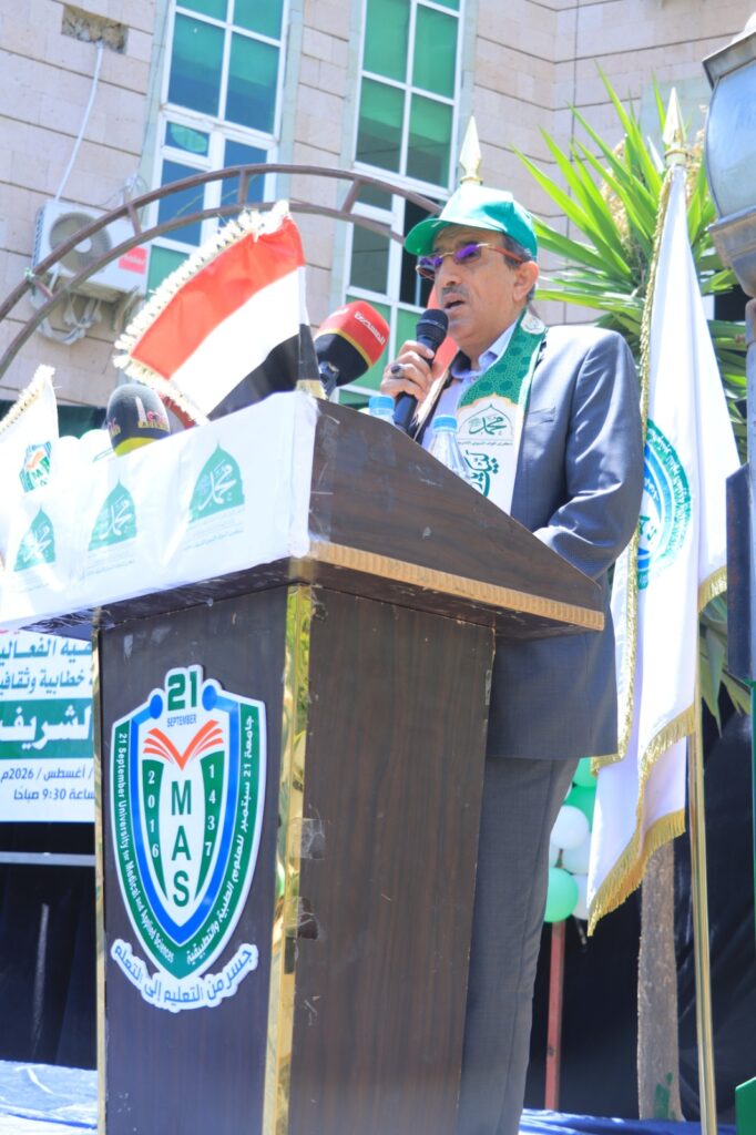 Man in a suit and green cap speaks into a microphone at a podium adorned with MAS banners, with flags in the background.
