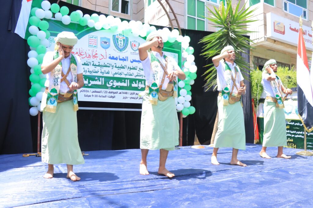 Four men in traditional Yemeni attire perform a synchronized salute on a blue stage at an outdoor cultural event, with a green-and-white balloon arch and an Arabic banner in the background.
