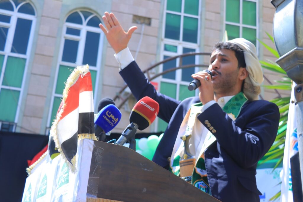 Man in a dark suit with a white headscarf speaks into a microphone at a podium, raising his left hand during an outdoor event with Yemeni flags and reporters nearby.