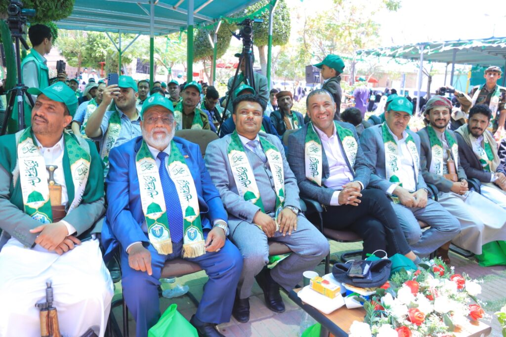 Group of men in suits wearing green ceremonial scarves and caps, seated in a row at an outdoor event with cameras and onlookers nearby.