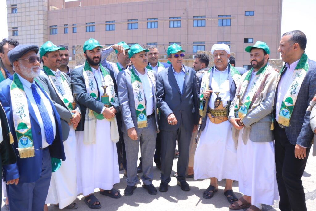 Group of men in traditional white robes and suits, wearing green scarves and hats, some with ceremonial swords, standing outside a building.