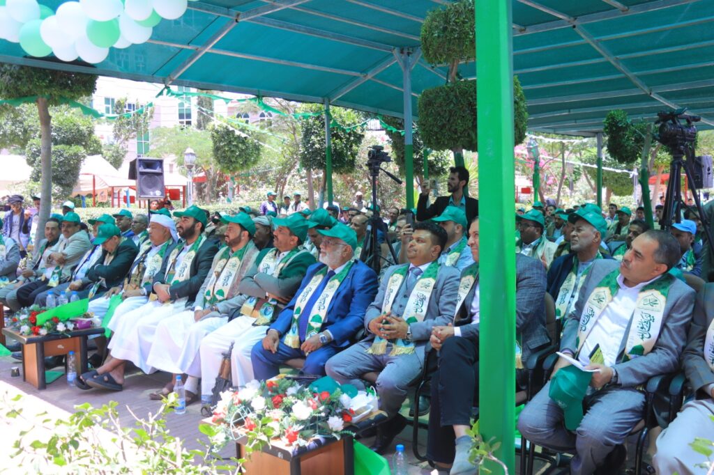 Group of men in green hats and sashes seated under a turquoise canopy at an outdoor ceremony