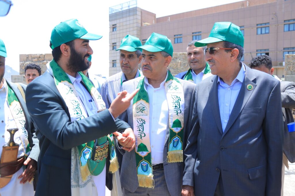 Group of men in green caps and decorative sashes talking outdoors, with a modern building in the background.