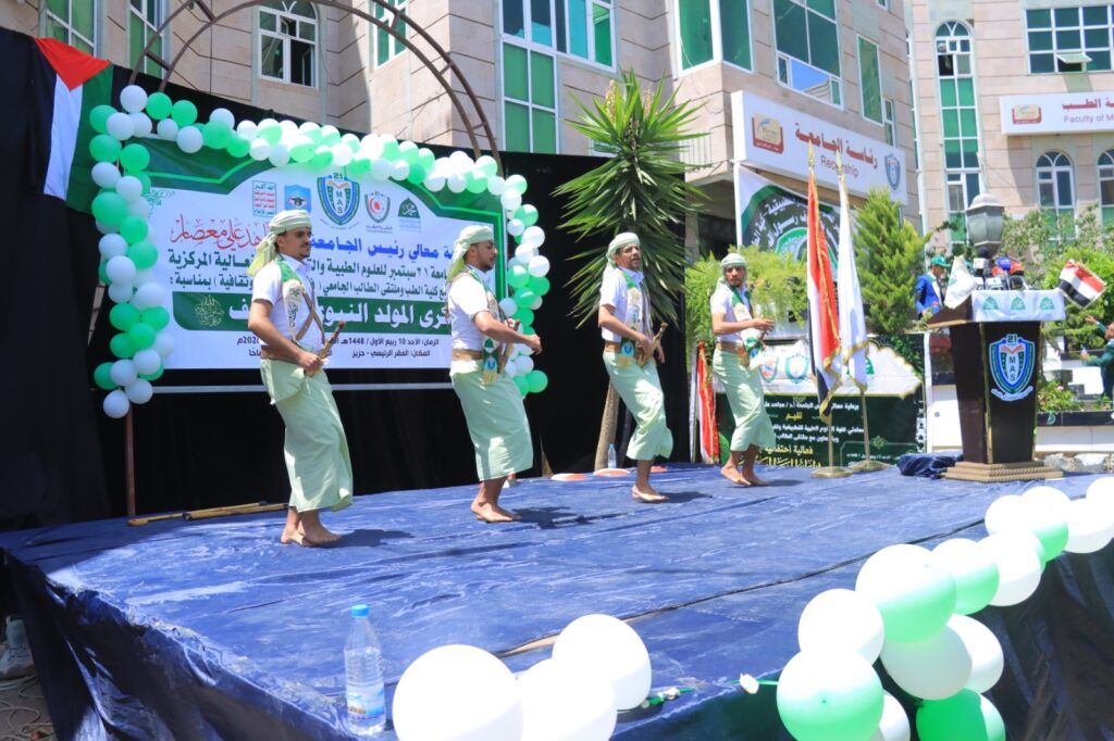 Four men in light-green outfits perform a dance on a blue stage framed by green-and-white balloons at a university event onlookers nearby.