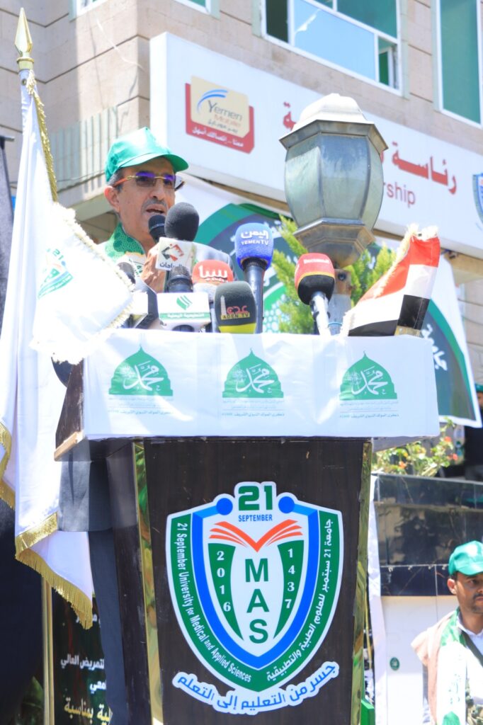 Man wearing a green cap speaks at a podium with multiple microphones during a public event; MAS university emblem on the podium banner behind him.