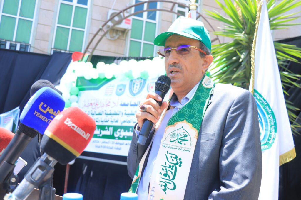 Man in a green cap and scarf speaks into a handheld microphone at a press event, with banners and reporters in the background.