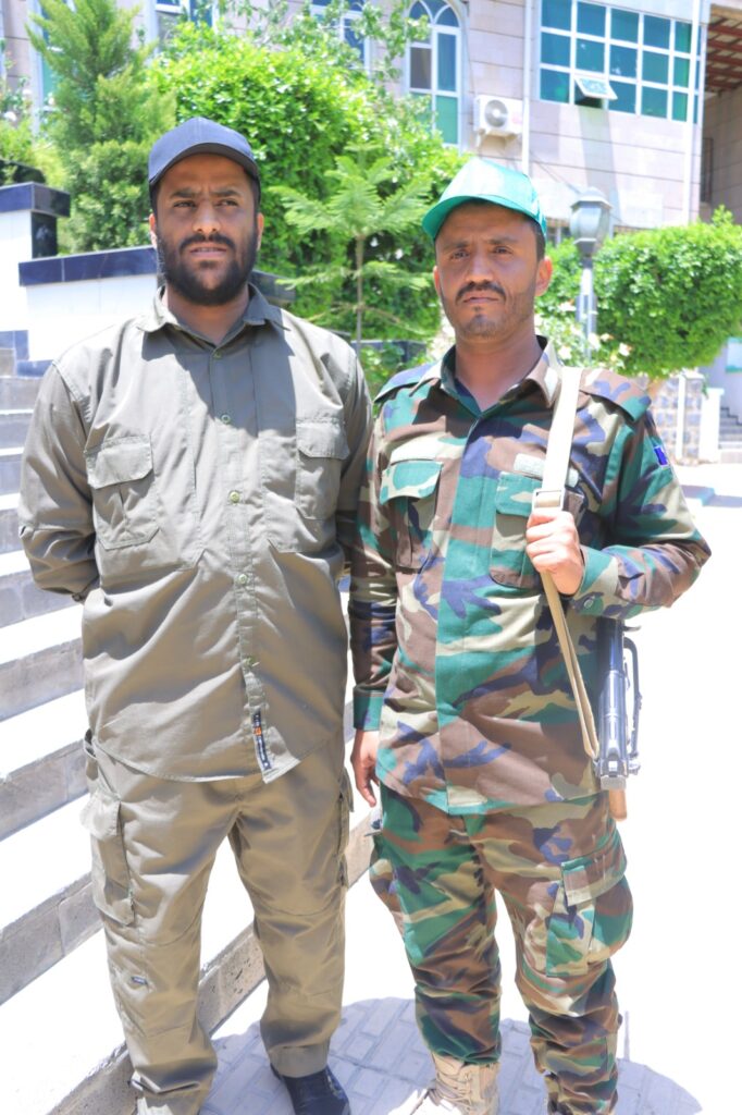 Two men in uniform posing outdoors side by side; one in olive-tan gear, the other in woodland camouflage with a teal cap, with greenery and a building behind them.