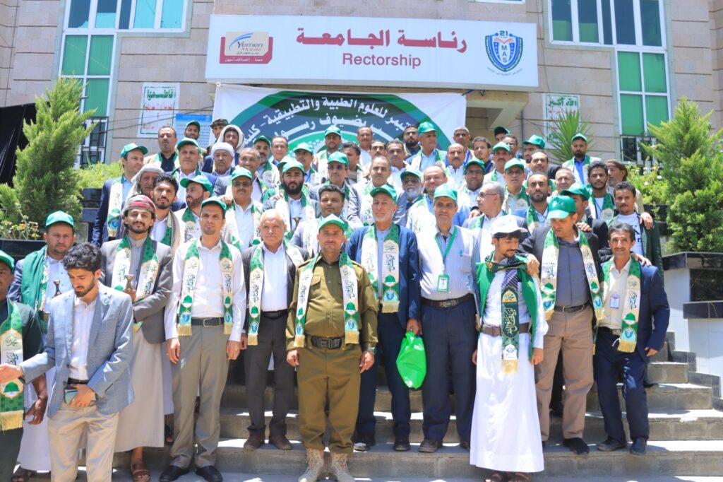 Group of men wearing green scarves and caps posing on steps in front of a university building during an event.