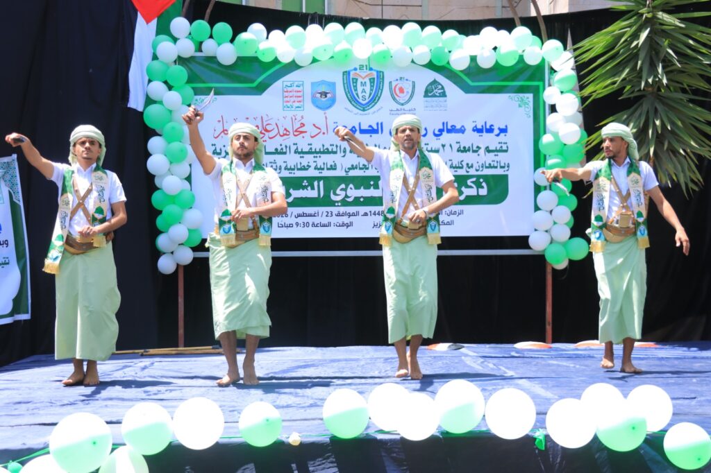 Four men in white shirts and green traditional outfits perform a dance on a decorated stage with green‑and‑white balloons and an Arabic banner behind them.