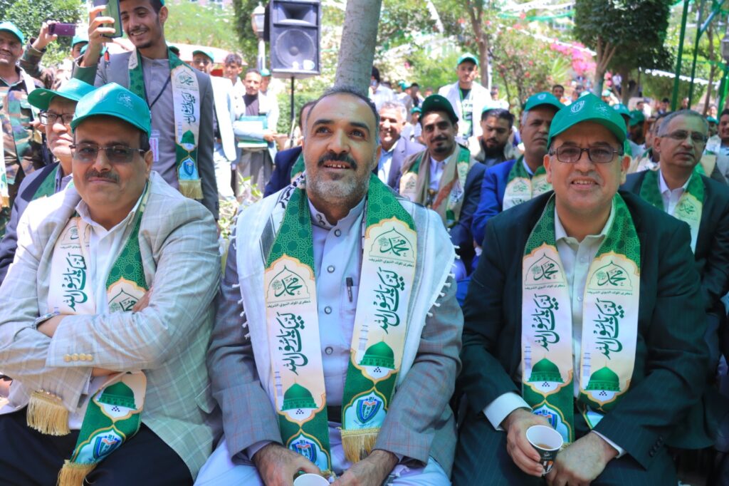 Group of men in suits wearing green commemorative scarves at an outdoor ceremony, smiling in a park setting.