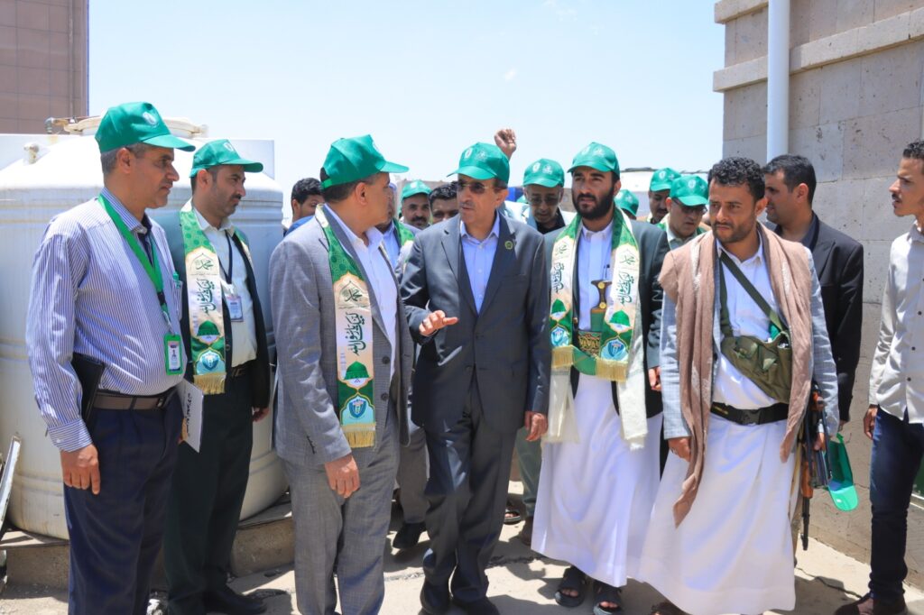 Group of men wearing green hats and ceremonial sashes outdoors, possibly attending a site visit or event together.
