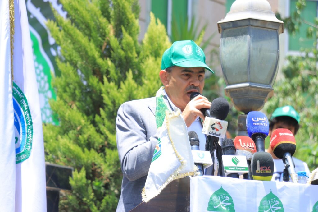 Man in a gray suit and green cap speaks into multiple microphones at a podium outdoors, with banners in the background.