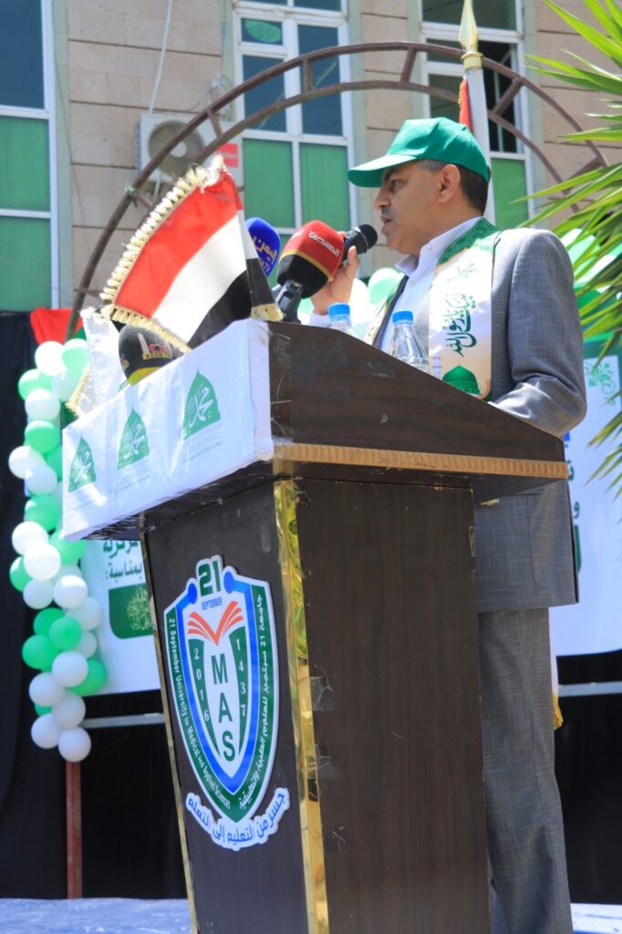 Man at a podium giving a speech, wearing a green cap and white scarf, with microphones and a flag beside him at an outdoor event.