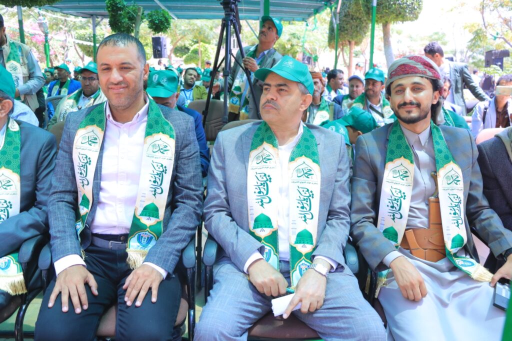 Three men in suits wear green ceremonial sashes at an outdoor event, seated in a crowd.