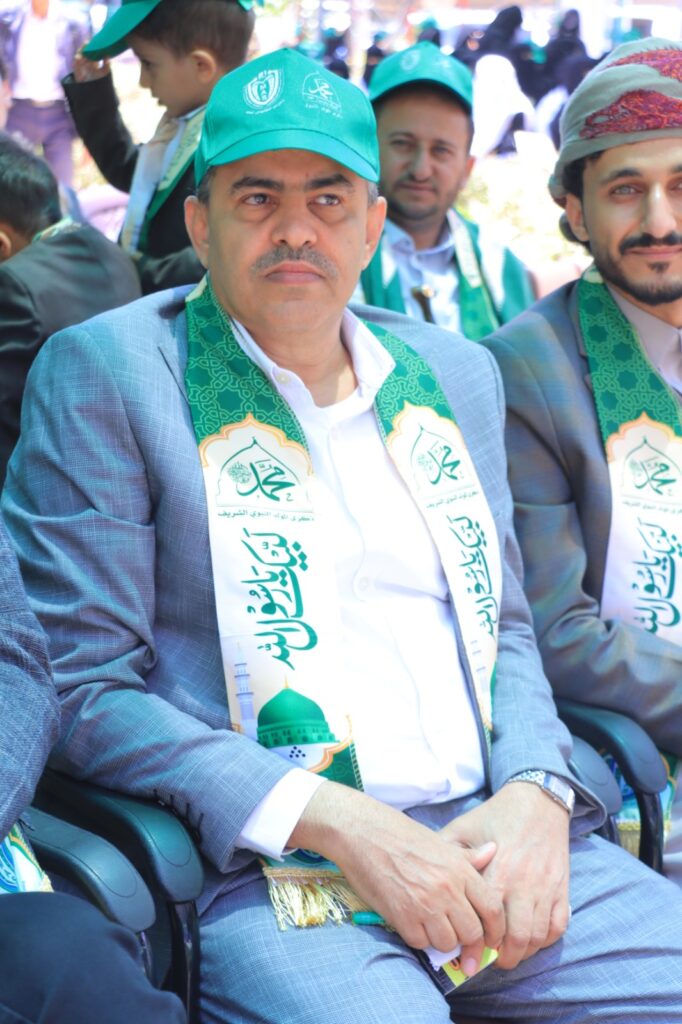 Middle-aged man in a blue suit seated outdoors, wearing a green cap and ceremonial green-and-white sash with Arabic text and a mosque illustration.