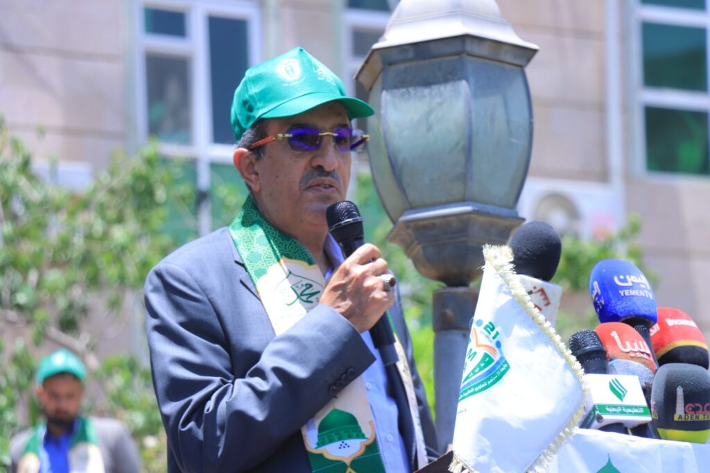Man in a blue suit and green cap speaks into a microphone at an outdoor press event with multiple microphones nearby, including news groups.