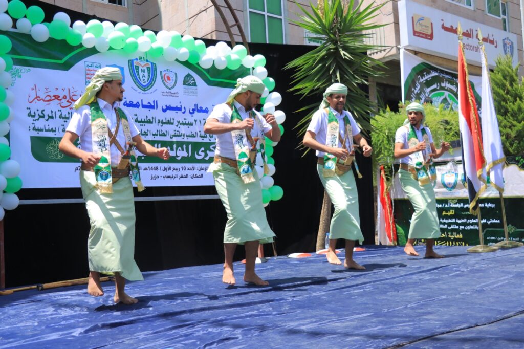 Four men in traditional attire perform a dance on a blue stage, with green sashes and headbands, in front of a banner and green-white balloons.