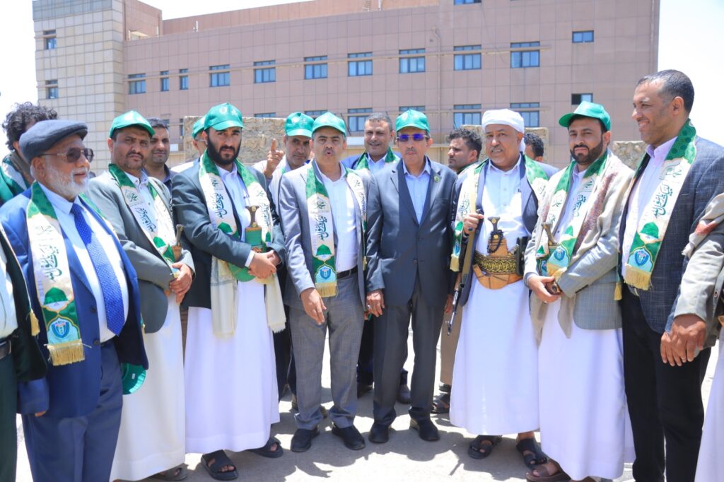 Group of men in traditional robes with green sashes posing for a photo outside a modern beige building.